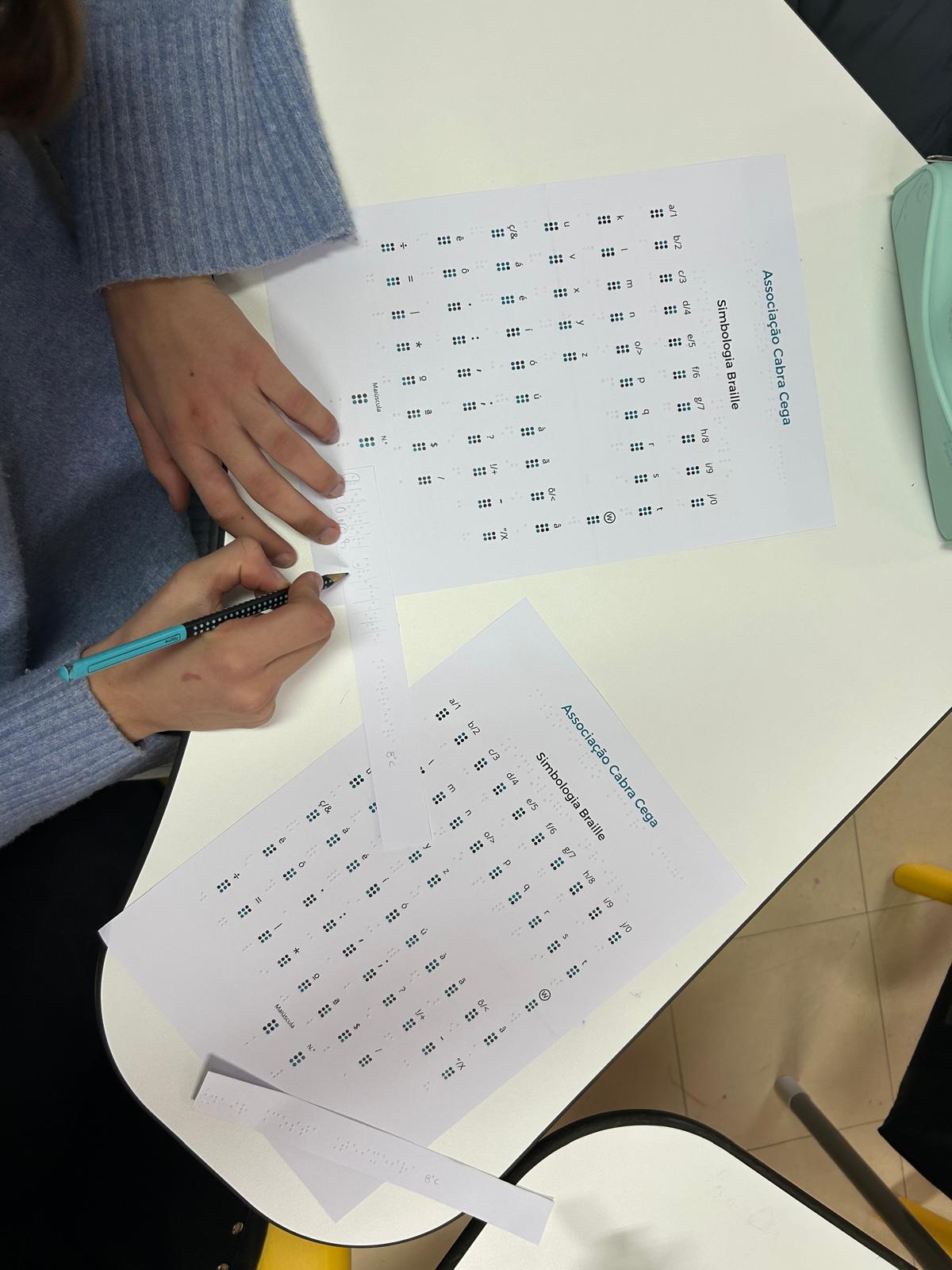 Uma mesa branca com folhas impressas de um alfabeto Braille e mãos de uma pessoa a usar uma tira de papel em Braile. Veem-se três folhas A4 com um alfabeto Braille. No topo de uma das folhas dá para ler “Associação Cabra Cega” e, abaixo, “Simbologia Braille”. A pessoa, com camisola de malha azul clara, segura a tira de papel e parece estar a decifrar uma mensagem. À direita há um objeto verde-claro (parece um estojo). No chão, no canto inferior direito, aparece parte de uma perna de cadeira amarela e o piso em azulejo.